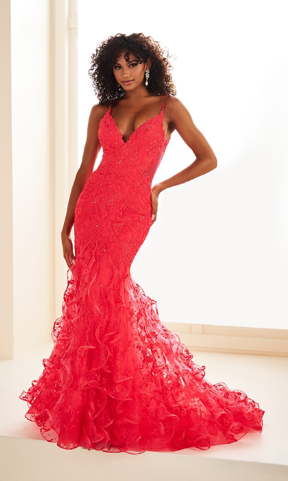 A woman in a fitted, floor-length bright red Ellie Wilde EW36041 prom dress with a v-neckline and ruffled hem stands against a light background.